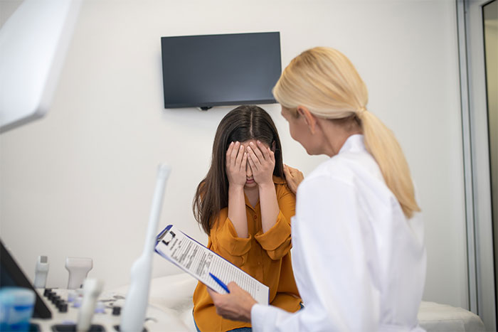 A woman in a doctor&rsquo;s office, holding a clipboard, sits across from a girl covering her face, discussing daughter&rsquo;s disease.