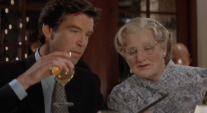 Two characters from a movie sit at a table, one holding a wine glass, sharing a moment possibly involving a long-standing joke.