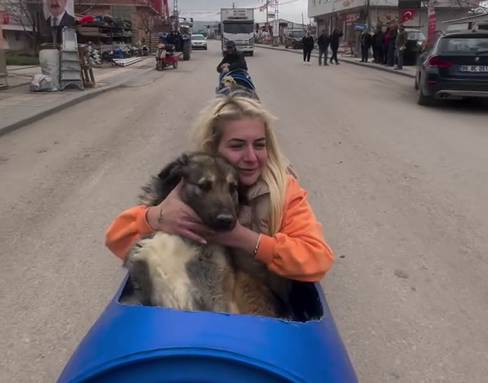 Woman riding a homemade dog train with a large dog, smiling as they travel down a street.