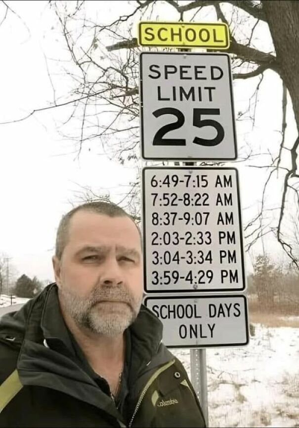 Man standing by a weird sign with unusual speed limit times near a school zone, in a snowy setting.