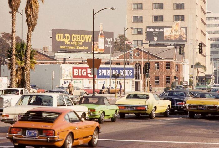 1970s West Coast street scene with vintage cars, billboards, and palm trees in urban landscape.