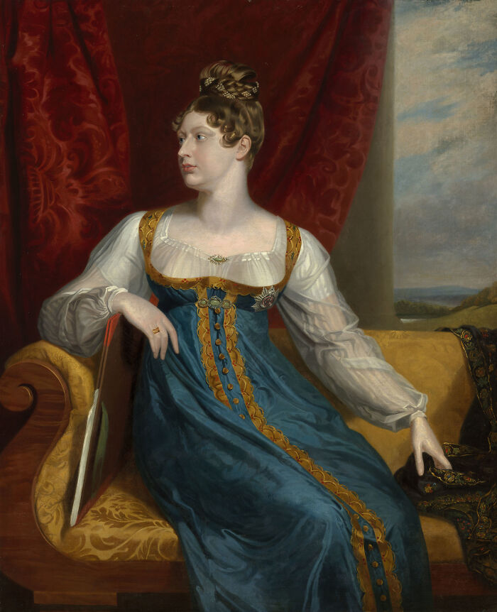 A woman in historical attire sitting on a yellow sofa with a red curtain in the background, symbolizing historical influence.