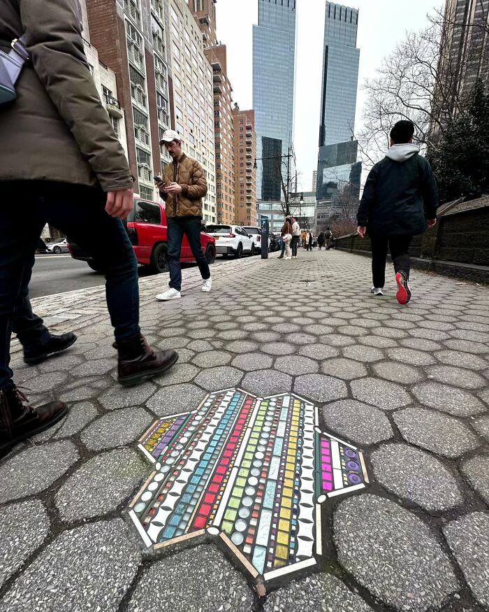 Vibrant mosaic art fills a c*****d sidewalk in a city, surrounded by pedestrians and tall buildings.