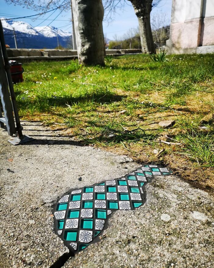 Vibrant mosaic art mends a c*****d sidewalk near a grassy area with trees and mountains in the background.