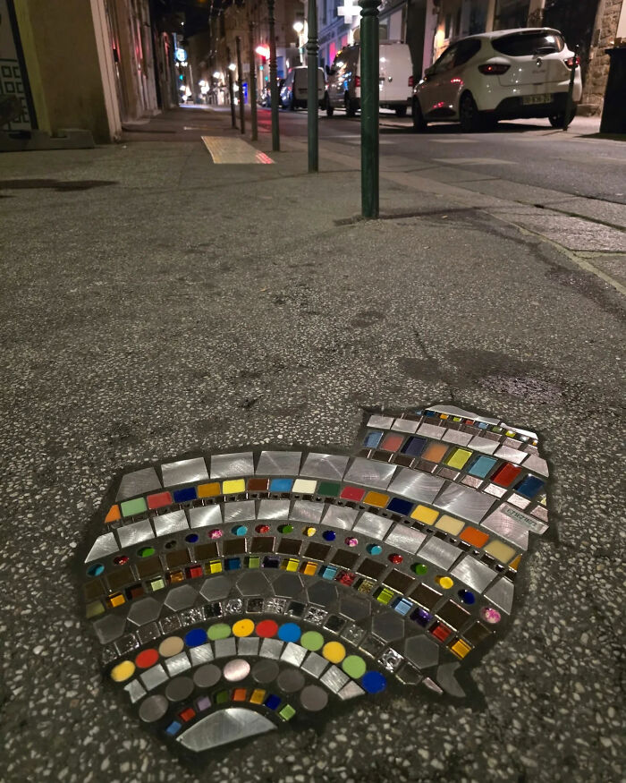 Vibrant mosaic art mending a c*****d sidewalk on a dimly lit street at night, with colorful tiles.