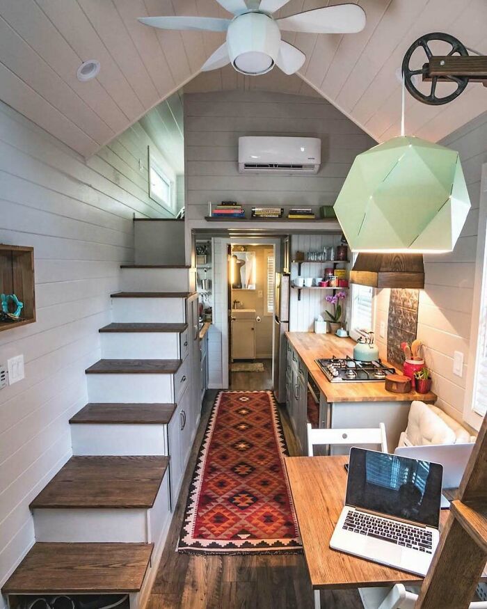 Cozy tiny home interior with wood accents, loft stairs, and modern kitchen featuring open shelving and a vibrant rug.