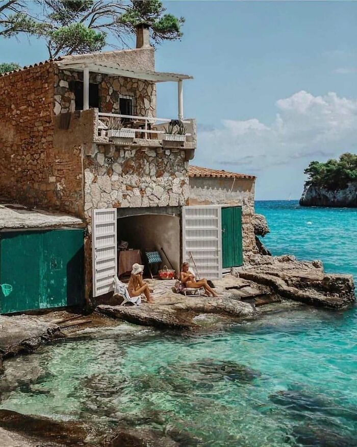 Scenic tiny home by the sea with two people relaxing outside, surrounded by turquoise water and stone architecture.