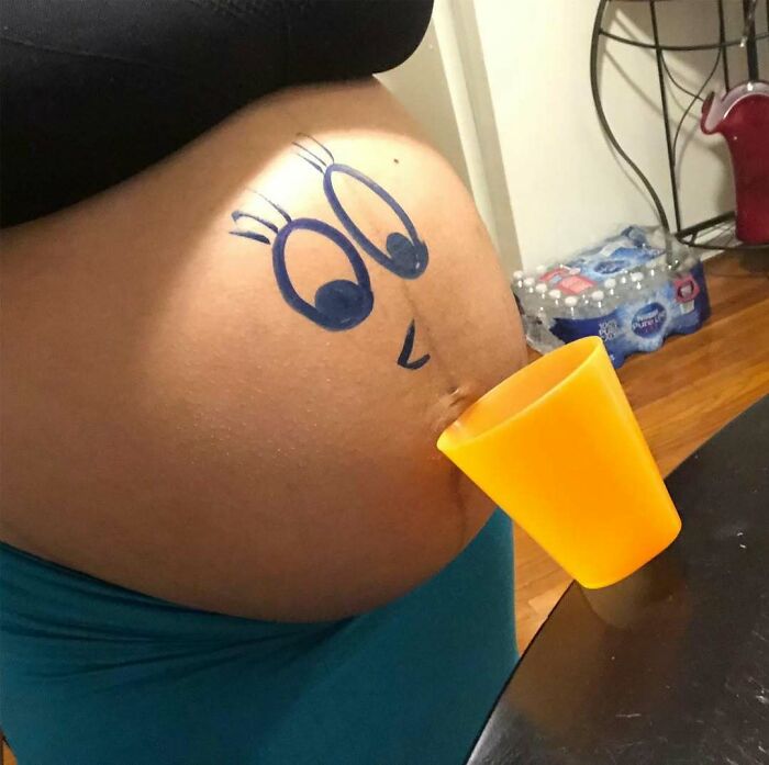 Pregnant belly with cartoon face drawn, holding an orange cup for a humorous pregnancy situation.