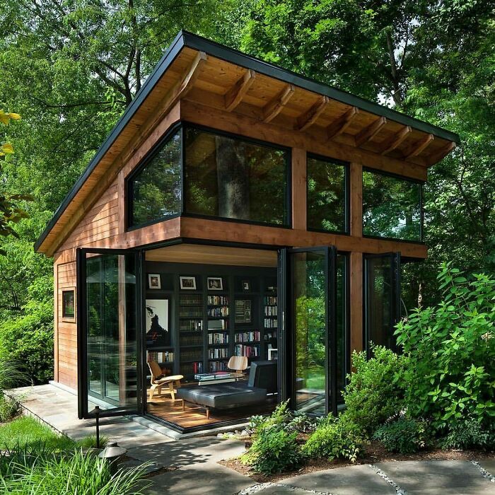 Tiny home surrounded by lush greenery, featuring large windows, bookshelves, and a cozy sitting area.