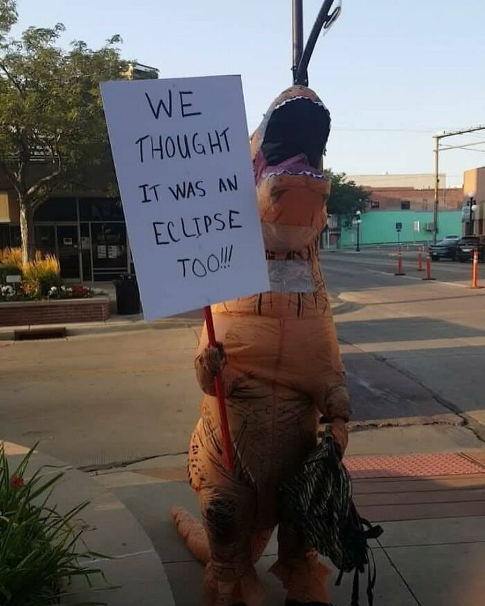 Person in dinosaur costume holding a sign about an eclipse, adding prehistoric humor to the street scene.