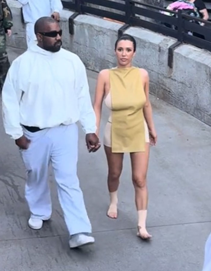 Bianca Censori walking barefoot in a unique dress, holding hands with a man in a casual white outfit outdoors.