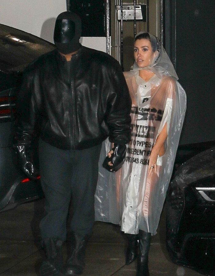 Bianca Censori in an edgy plastic raincoat and boots, accompanied by a masked man in black leather jacket at night.