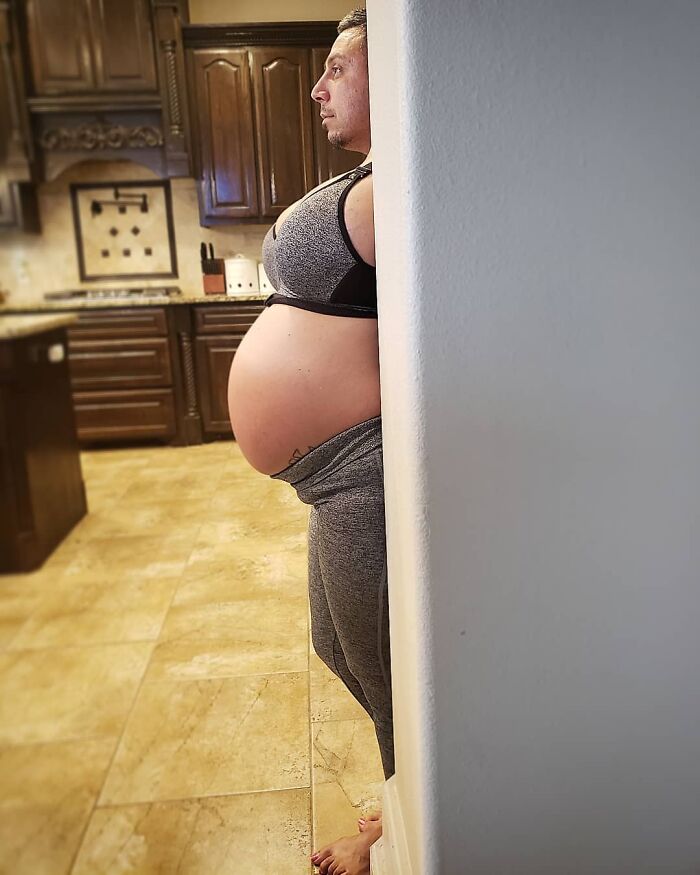 Person wearing a pregnancy belly suit, humorously standing in a kitchen.