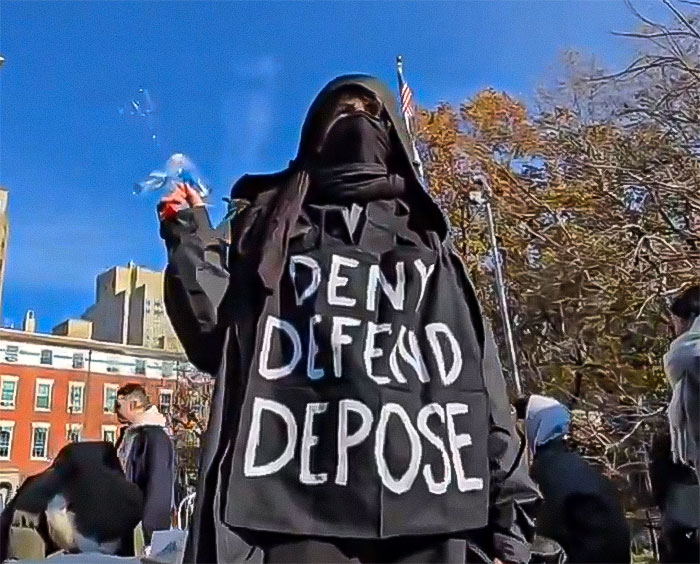 Protester in dark clothing at a park, holding a bubble maker, with "Deny Defend Depose" on the shirt.