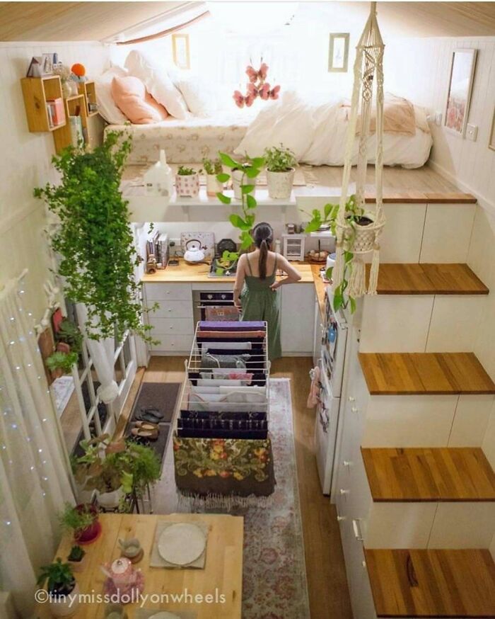 Tiny home interior with a woman in a green dress, surrounded by plants, kitchen area, and loft bed above.