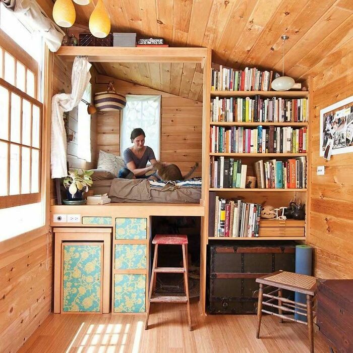 Cozy tiny home interior with wooden walls, woman on elevated bed, bookshelf, stool, and vintage decor.