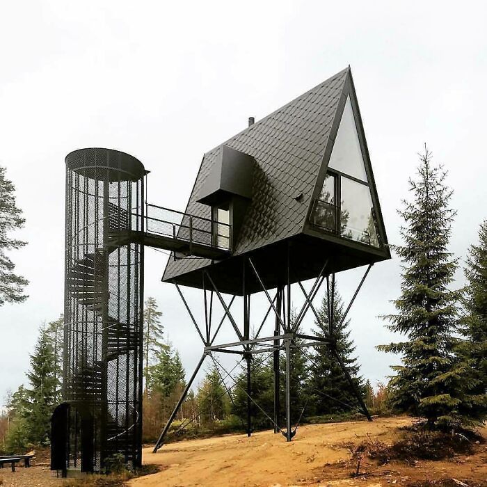A modern tiny home elevated on stilts with a spiral staircase in a forest setting.