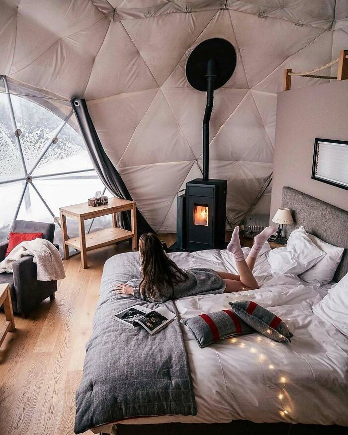 Cozy interior of a tiny home with a woman reading on a bed near a window and wood stove.