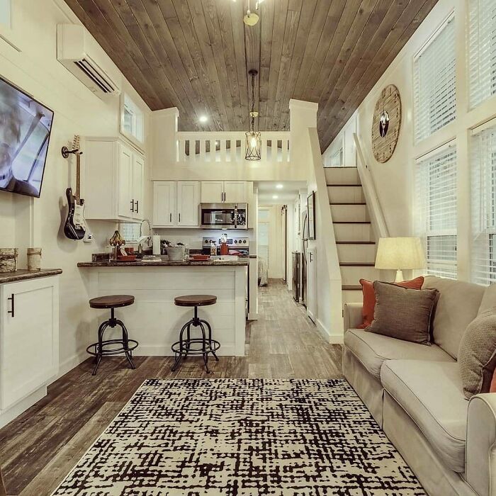 Cozy interior of a tiny home with wooden ceiling, compact kitchen, and stairs leading to a loft, featuring modern decor.