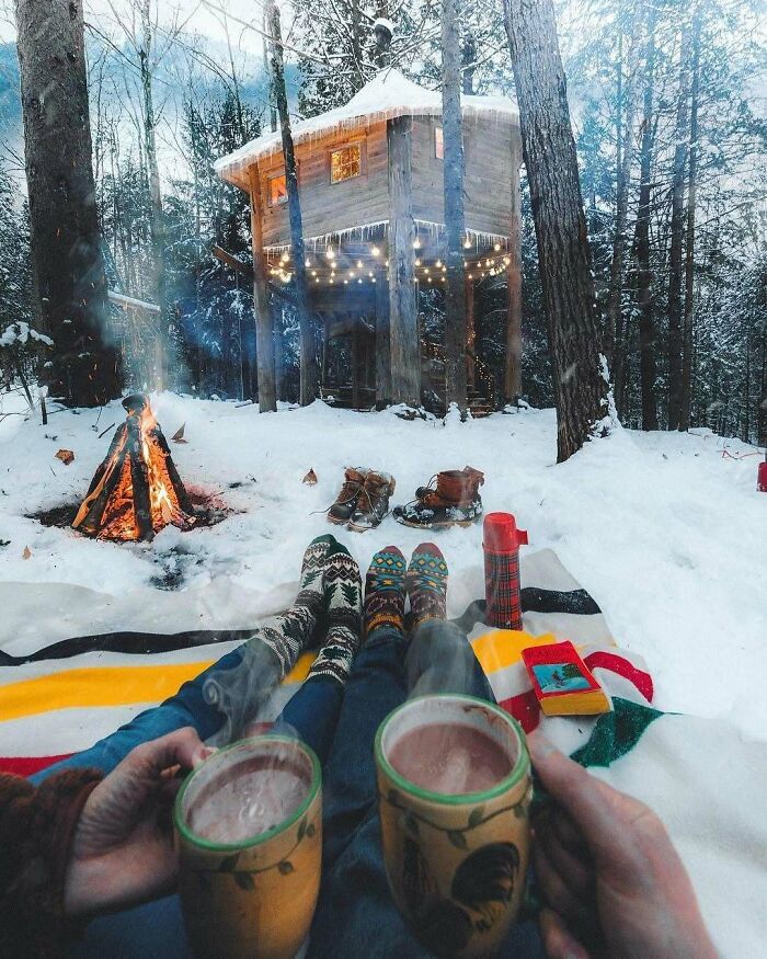 Cozy winter scene with steaming mugs in front of a tiny home treehouse, surrounded by snow and a warm campfire.