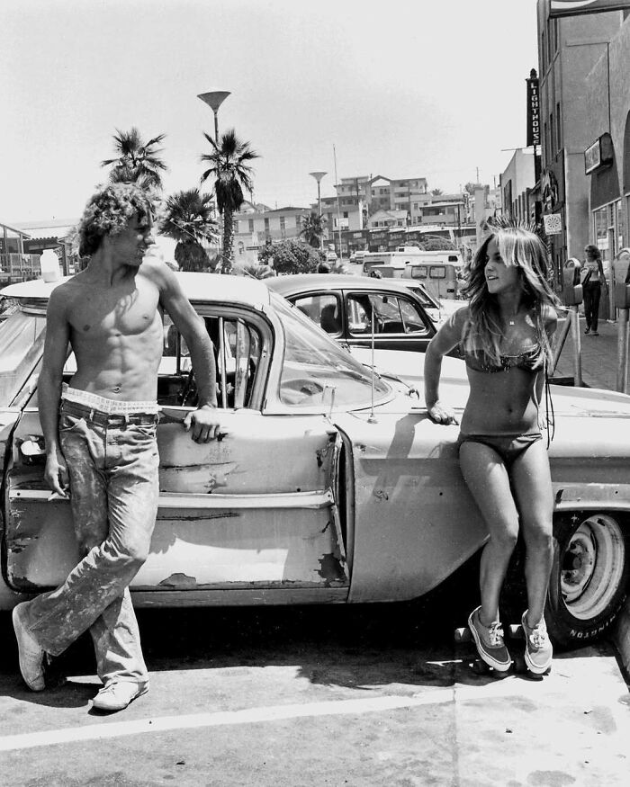 Two people in 1970s West Coast fashion by a classic car, with palm trees and buildings in the background.