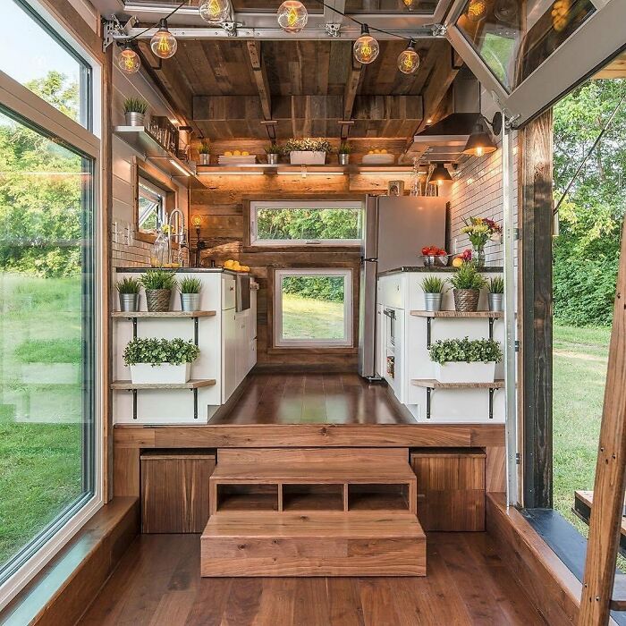 Interior view of a charming tiny home featuring wooden floors, white cabinets, and large windows overlooking greenery.