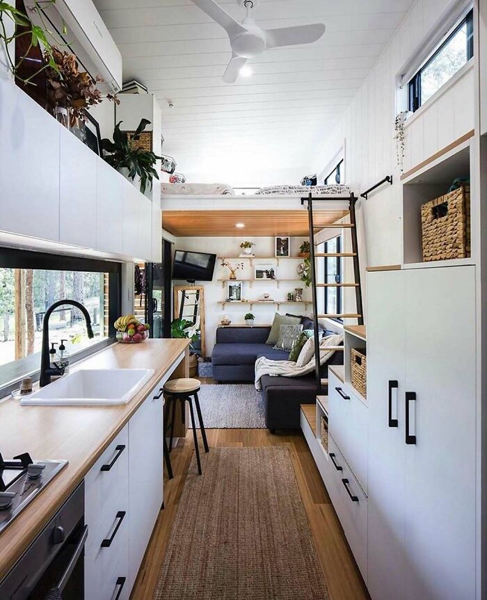 Modern tiny home interior with loft bed, cozy living area, and compact kitchen featuring sleek white cabinets and wood accents.
