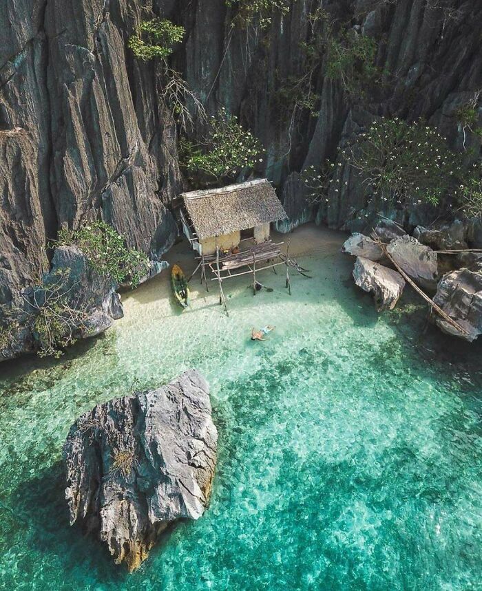 Tiny home beside a turquoise lagoon surrounded by rocky cliffs, with a kayak and a person swimming nearby.