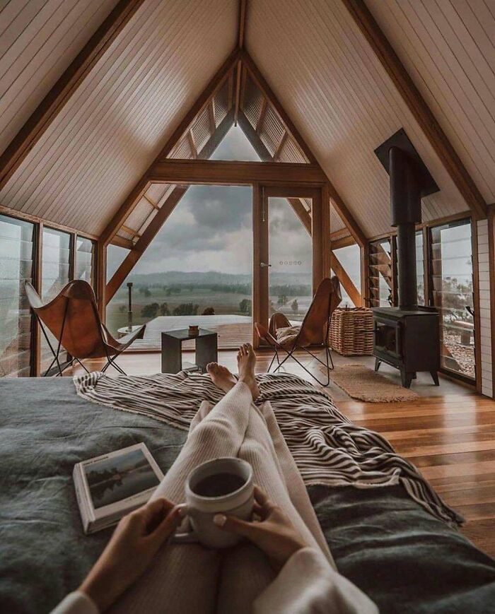 Cozy interior of a tiny home with wooden beams, large windows, and a person holding a cup of coffee.