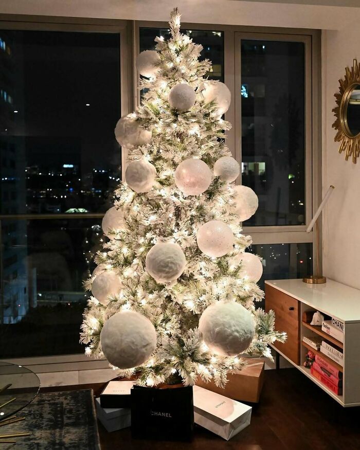 Elegant Christmas tree with large white baubles, lit up in a modern living room setting at night.