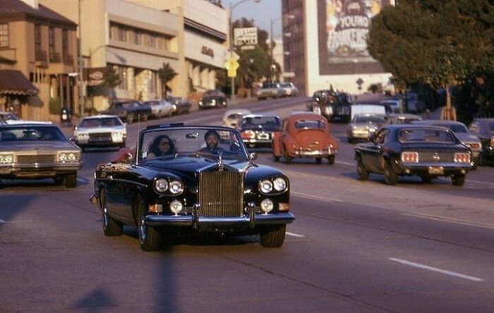 Vintage 1970s West Coast street scene with classic cars, including a black convertible, on a sunny day.