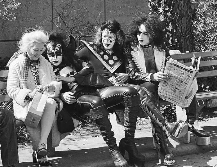 Historical photo of rock band members in costume sitting on a park bench with an elderly woman, blending past and future.