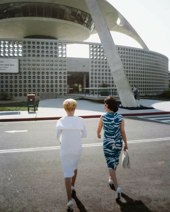 Two women in 1970s West Coast fashion, walking near a futuristic building with a lattice facade.