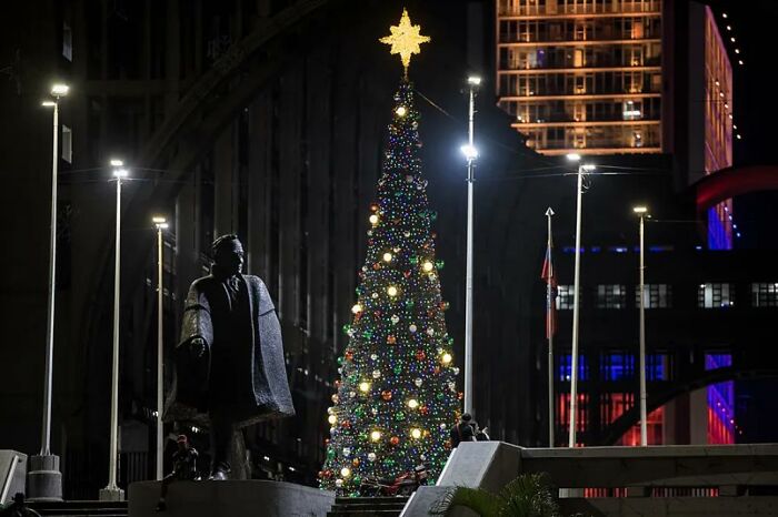 Christmas tree adorned with lights next to a statue in a city plaza at night.