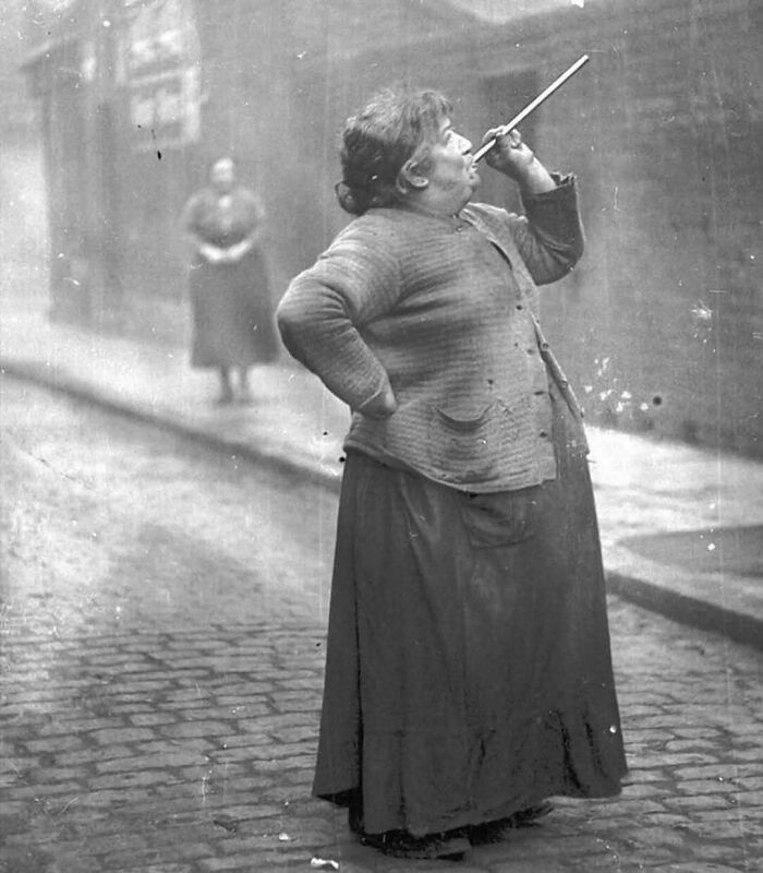 Woman in historical attire blowing a long pipe on a cobblestone street, showcasing interesting history pics.