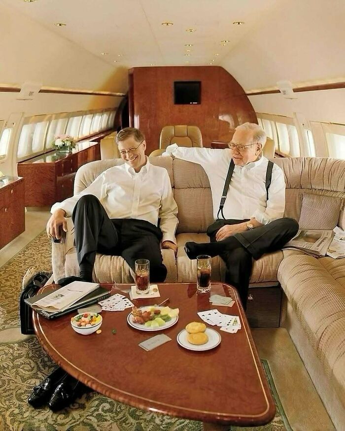 Two men in an aircraft lounge discussing investments, with snacks and playing cards on the table.