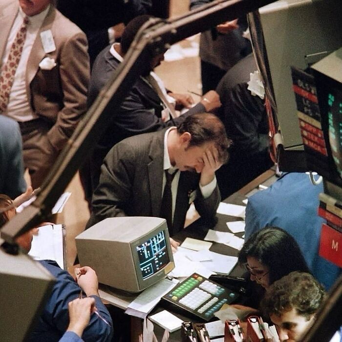 Trader at Wall Street desk with vintage computer, looking stressed. Investment professional surrounded by busy colleagues.