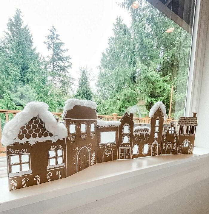 DIY Christmas village decoration on windowsill with snowy rooftops, framed by lush green trees outside.