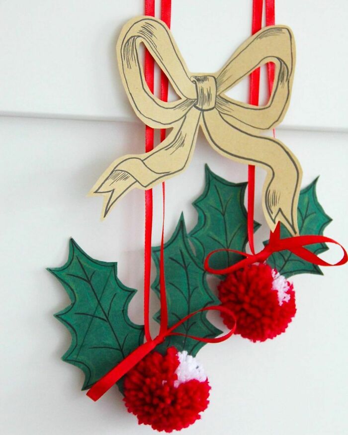 DIY Christmas decorations with holly leaves, red pom-poms, and a decorative bow hanging on a wall.