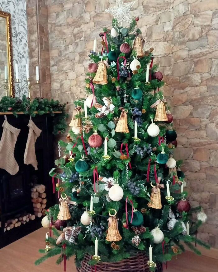 Christmas tree decorated with natural ornaments, wicker bells, and candles, set against a stone wall background.