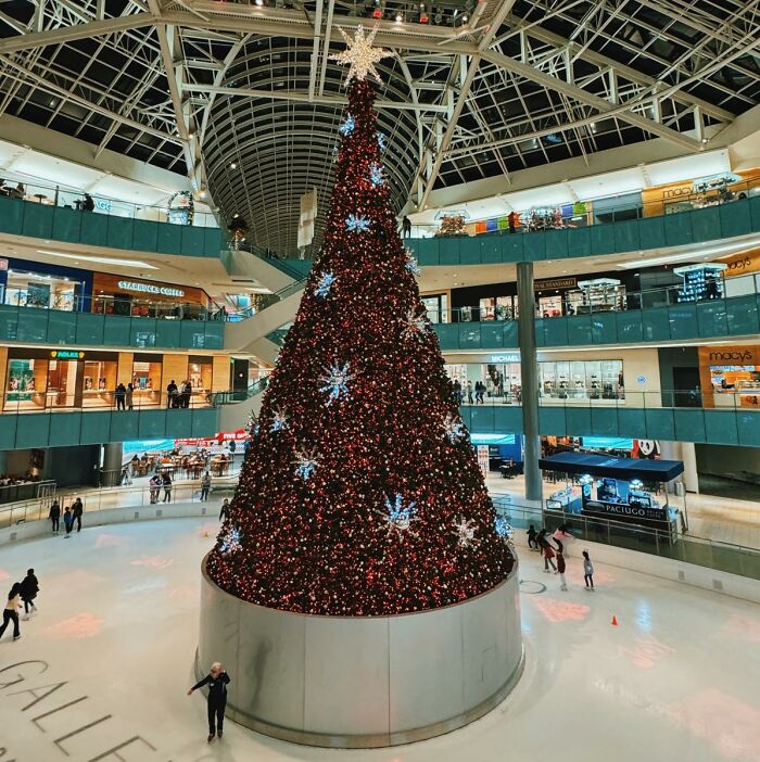 Tall Christmas tree in a shopping mall, surrounded by shoppers on an ice rink, showcasing festive decorations globally.