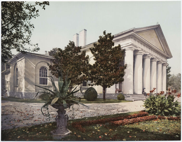 Historic color photo of a neoclassical building with columns and surrounding garden.
