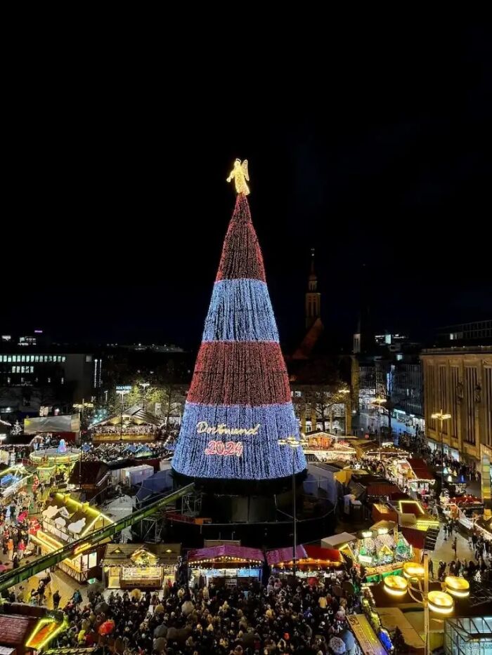 Christmas tree in Dortmund decorated with lights and a glowing angel, surrounded by festive market stalls at night.