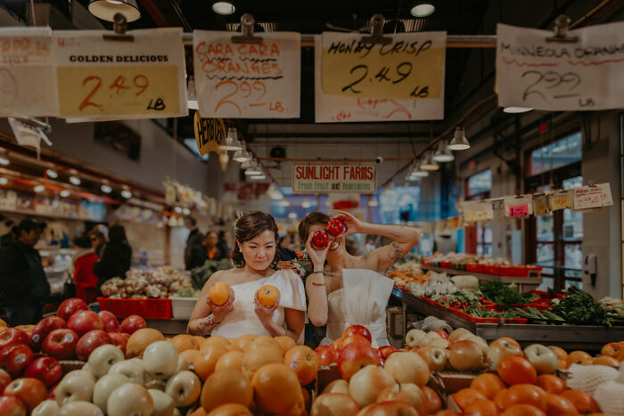 Brides at a market, surrounded by apples and oranges, capturing a unique wedding photo moment.