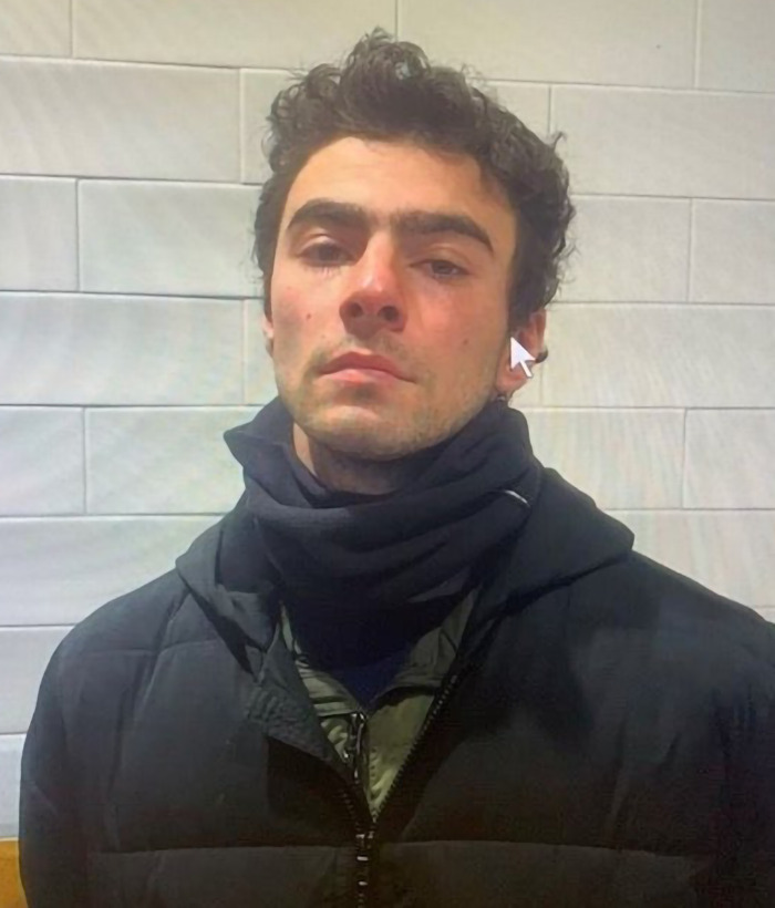 Luigi Mangione in a black jacket and scarf standing against a tiled wall.