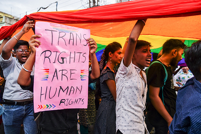 People holding a pink sign reading "Trans Rights Are Human Rights" under a rainbow flag at a protest.