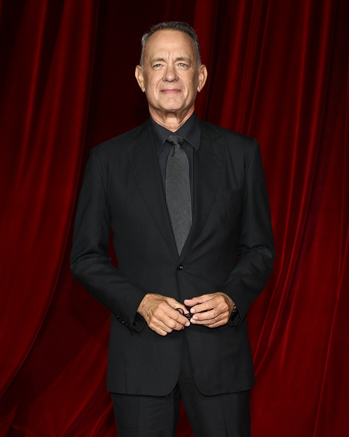 Tom Hanks in a black suit standing against a red curtain backdrop.