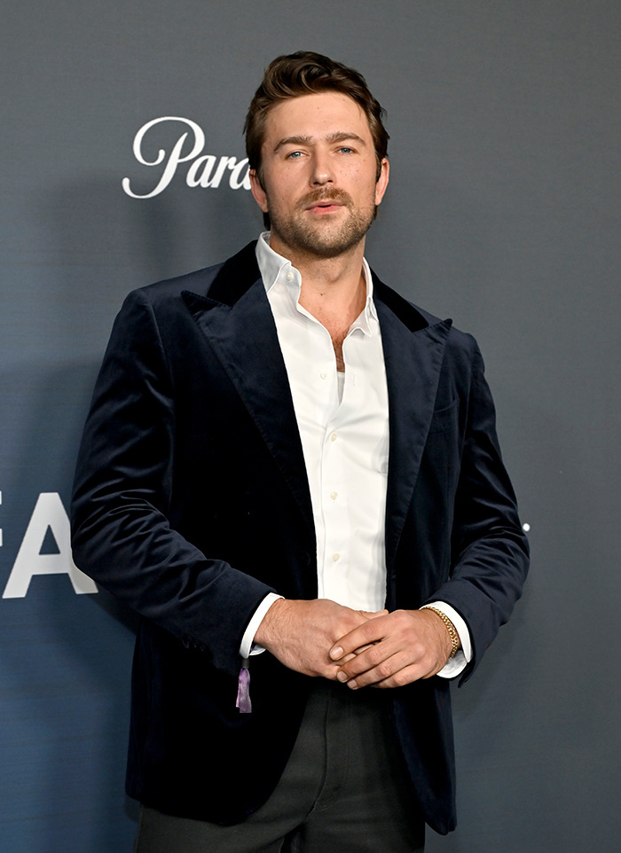Brandon Sklenar wearing a velvet jacket, stands confidently at an event backdrop. Brandon Sklenar wearing a velvet jacket, stands confidently at an event backdrop.