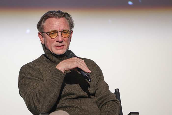 Daniel Craig in a brown sweater and yellow glasses speaking into a microphone at a film event.