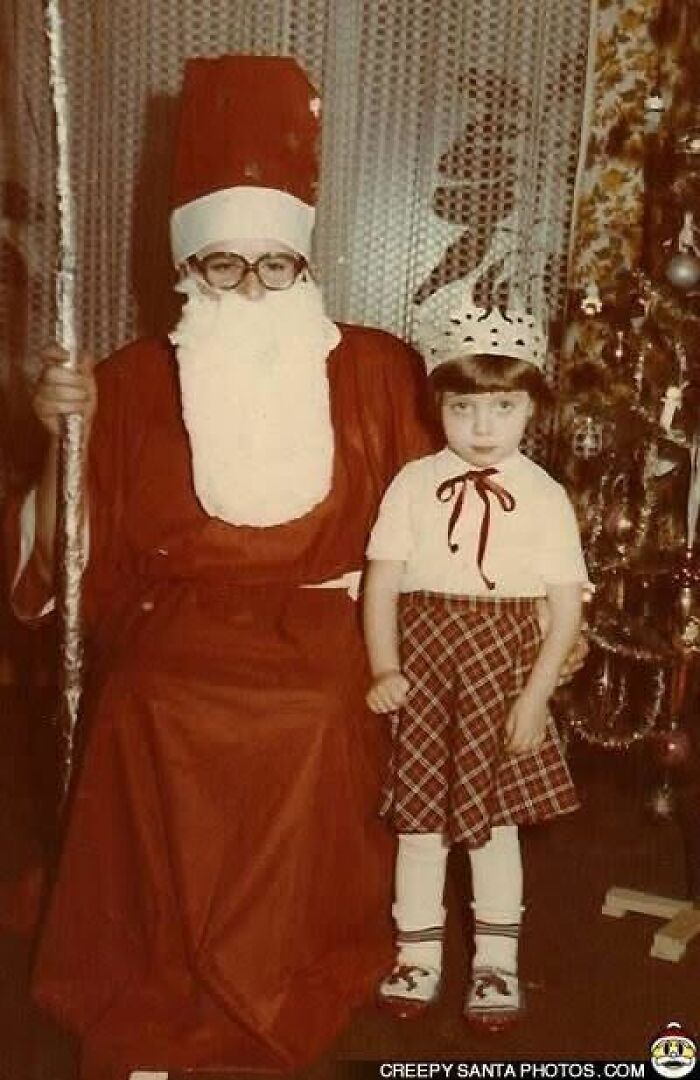 Child stands next to a creepy Santa in vintage attire with a decorated Christmas tree in the background.